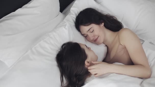 Pareja de lesbianas abrazándose y sonriendo mientras yacen juntas en la cama en casa. Jóvenes lesbianas besos y abrazos después de despertar — Vídeo de stock