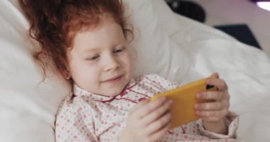 Komik kızıl saçlı küçük kız ilgi ile modern sarı akıllı telefon video izlerken yatakta yatan.