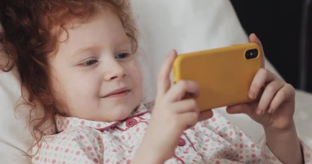 Divertida pelirroja niña mintiendo en la cama viendo vídeo en el teléfono inteligente amarillo moderno con interés . — Vídeos de Stock