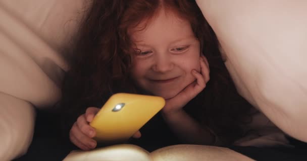 Μια μικρή κοκκινομάλλα που διαβάζει βιβλίο με ενδιαφέρον για το κρεβάτι του στο σπίτι. Κορίτσι χρησιμοποιώντας φακό του smartphone για τις σελίδες φωτισμού. — Αρχείο Βίντεο