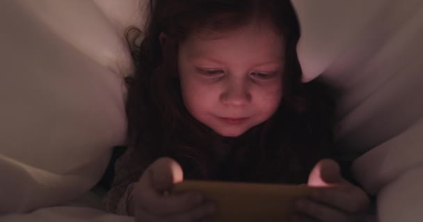Pelirroja niña viendo vídeo en su teléfono inteligente amarillo por la noche. Se esconde debajo de la manta. . — Vídeos de Stock