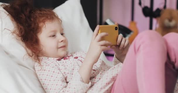 Divertida pelirroja niña mintiendo en la cama viendo vídeo en el teléfono inteligente amarillo moderno con interés . — Vídeos de Stock