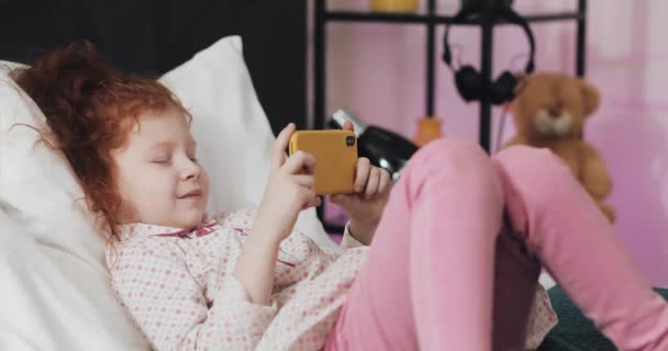 Menina ruiva engraçada deitada na cama assistindo vídeo no smartphone amarelo moderno com interesse . — Vídeo de Stock