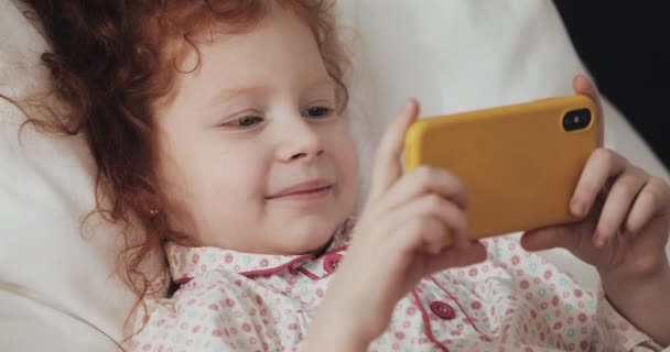 Забавная рыжая маленькая девочка лежит в постели и смотрит видео на современном желтом смартфоне с интересом . — стоковое видео