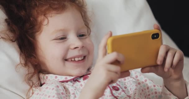 Divertente rossa bambina sdraiata sul letto a guardare video al moderno smartphone giallo con interesse . — Video Stock