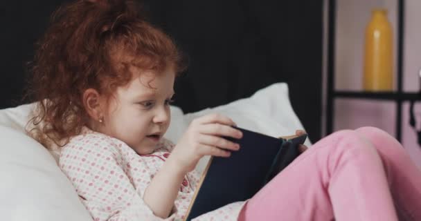 Divertida pelirroja niña leyendo un libro en la cama en su dormitorio . — Vídeos de Stock