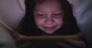 Kızıl saçlı küçük kız geceleri sarı akıllı telefonunda video izliyor. Battaniyenin altında saklanıyor..
