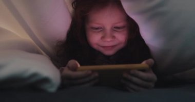 Kızıl saçlı küçük kız geceleri sarı akıllı telefonunda video izliyor. Battaniyenin altında saklanıyor..