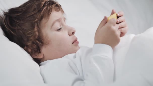 興味を持って現代の黄色のスマートフォンで漫画を見てベッドに横たわっている小さな男の子. — ストック動画