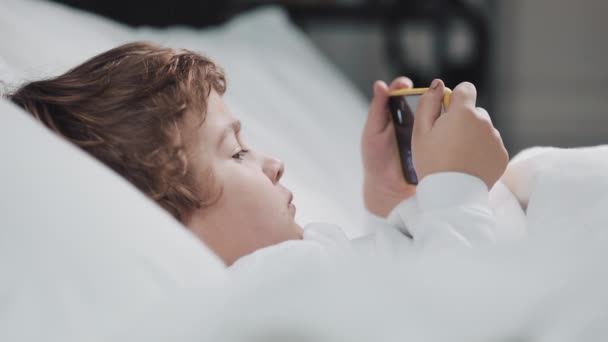 Mały chłopiec leżący w łóżku oglądając kreskówki na nowoczesnym żółtym smartfonie z zainteresowaniem. — Wideo stockowe