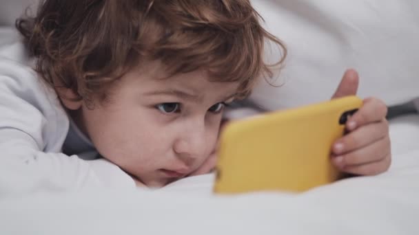 Niño tendido en la cama viendo dibujos animados en el teléfono inteligente amarillo moderno con interés . — Vídeo de stock