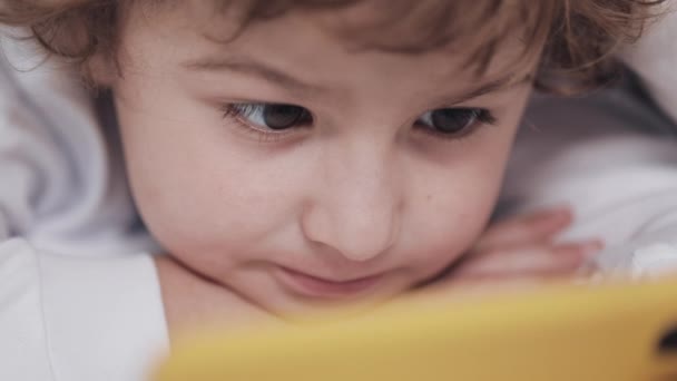 Menino deitado na cama assistindo desenhos animados no smartphone amarelo moderno com interesse . — Vídeo de Stock