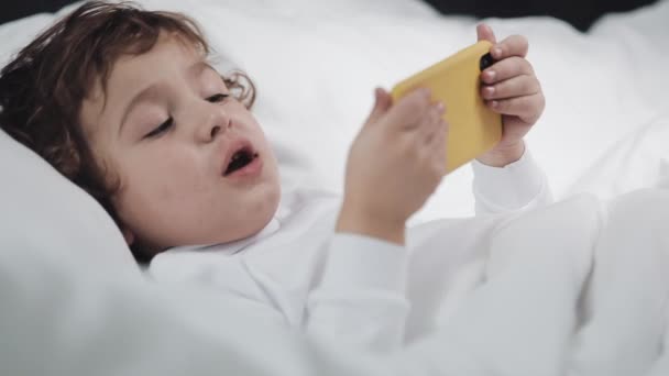 Niño con la tableta frotándose los ojos después de llorar. Se acuesta en la cama, sosteniendo un smartphone amarillo en sus manos . — Vídeos de Stock