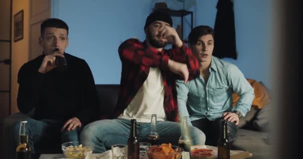 Tres amigos viendo el partido de fútbol en la televisión en casa y estando tristes porque su equipo pierde. El hombre lanza palomitas de maíz en la TV . — Vídeos de Stock