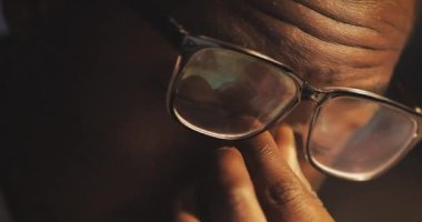 Bilgisayar üzerinde çalışmaktan bıkmış genç siyah adam gözlüklerini yukarı iter ve gözlerini ovuşturuyor. İşkolik, çok çalışıyor. Gece vakti. Yakın çekim.