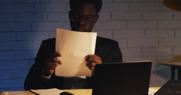 Cansado de trabajar con documentos, el joven negro se levanta las gafas y se frota los ojos. Workaholic, trabajando duro. Noche. De cerca. . — Vídeo de stock