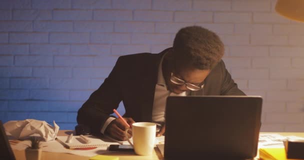 Joven hombre de negocios negro con computadora portátil y papeles trabajando en la oficina nocturna. Firma los documentos. Negocio, adicto al trabajo, concepto de plazo . — Vídeo de stock