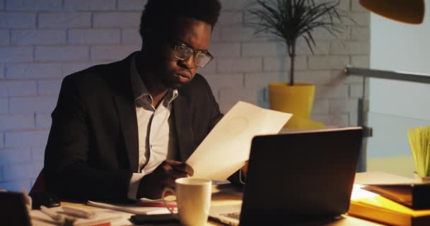 Tired young black businessman with laptop computer and papers working at night office. He working with documents. Business, workaholic, deadline concept. — Stock Video