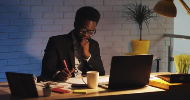 Joven hombre de negocios negro cansado con computadora portátil y papeles trabajando en la oficina nocturna. Trabaja con documentos. Negocio, adicto al trabajo, concepto de plazo . — Vídeos de Stock