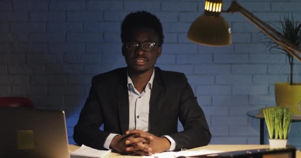 Portret van gelukkige jonge zwarte zakenman zittend aan de tafel op kantoor achtergrond 's nachts en kijken naar de camera. — Stockvideo