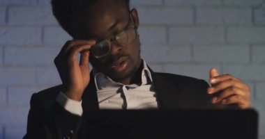 Bilgisayar üzerinde çalışmaktan bıkmış genç siyah adam gözlüklerini yukarı iter ve gözlerini ovuşturuyor. İşkolik, çok çalışıyor. Gece vakti. Yakın çekim.