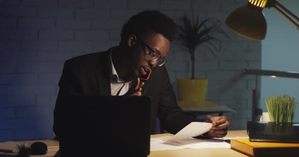 Jovem trabalhador de escritório afro-americano conversando ao telefone com o chefe, sentado no escritório moderno à noite, trabalhando duro com laptop. Ele está estressado e zangado . — Vídeo de Stock