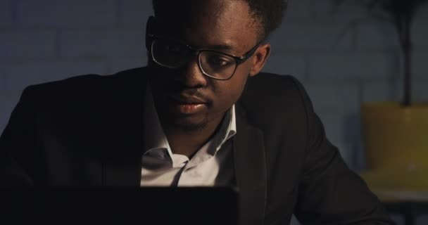 Hombre de negocios afroamericano concentrado en anteojos delante de la computadora portátil con reflejo de la exhibición en anteojos. Joven freelancer masculino trabajando de noche. De cerca. . — Vídeos de Stock