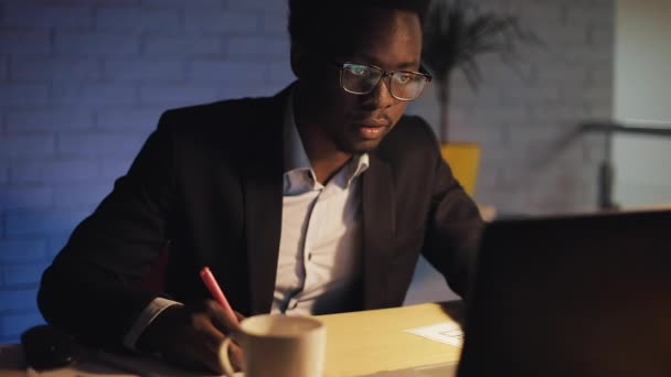年轻的黑人商人带着手提电脑和文件在夜间工作。 他在文件上签字，喝咖啡。 业务、工作狂、截止日期概念. — 图库视频影像