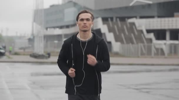 Jovem atraente correndo ao ar livre usando fones de ouvido câmera lenta. Ele está a olhar para a câmara. Tempo chuvoso. Exercício cardiológico. Estilo de vida saudável - execução . — Vídeo de Stock