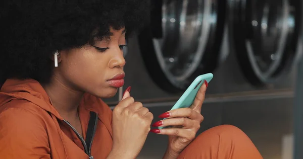 Çamaşırhanede akıllı telefon kullanan çekici genç kadın. Kablosuz kulaklık takıyor ve içeride şarkı söylüyor..