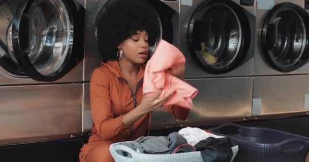 African American młoda kobieta sortowania prania w koszyku ubrania sortowania przed mycia siedzi w samoobsługowej pralni publicznej. — Wideo stockowe