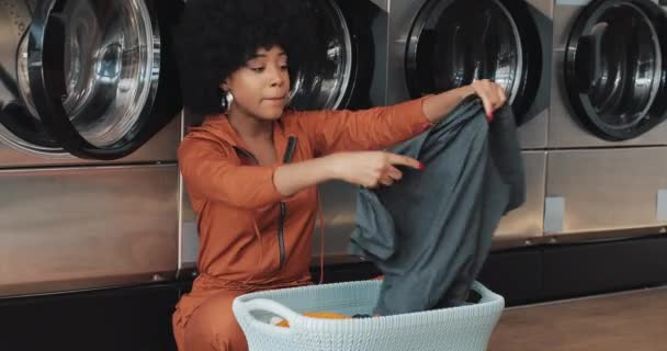 Jovem afro-americana classificando roupa na cesta classificando roupas antes de lavar sentado na lavanderia pública self-service . — Vídeo de Stock