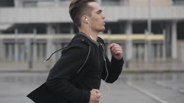 Atractivo joven corriendo al aire libre con auriculares en cámara lenta. Clima lluvioso. Entrenamiento de ejercicio cardiovascular. Estilo de vida saludable - correr . — Vídeos de Stock