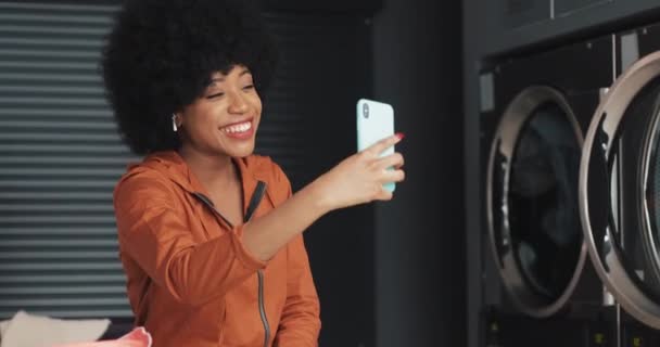 コインランドリーでビデオチャットを持っている幸せな魅力的なアフリカ系アメリカ人の若い女性。セルフサービスの公共ランドリー. — ストック動画