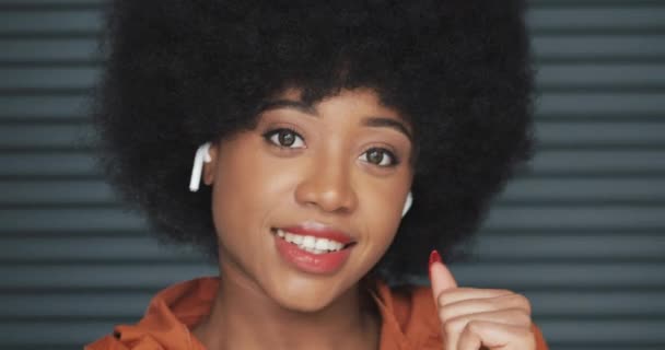 Retrato de una joven afroamericana feliz con auriculares, escuche música y baile divertido en la cámara. Persianas horizontales de fondo. Primer plano . — Vídeos de Stock