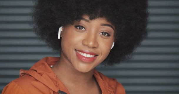 Portrait of young happy African American woman wearing earphones looking into the camera. Horizontal roller blinds background. Close-up. — Stock Video