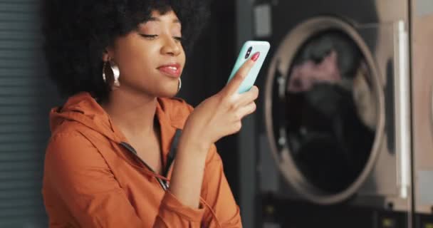 Mujer joven sonriente usando teléfono inteligente en la lavandería. Autoservicio de lavandería pública . — Vídeo de stock