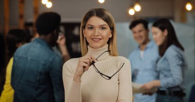 Ofis çalışma alanında duran kameraya bakan şık gözlüklü genç başarılı iş kadınının portresi. Bulanık arka plan üzerinde çalışan insanlar.