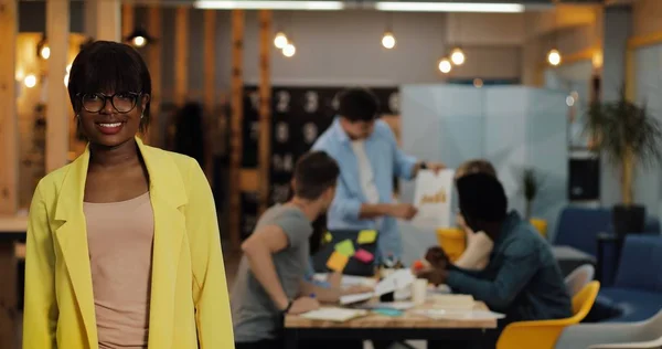 Afro-Amerikalı iş kadını modern ofiste ayakta, gülümseyerek ve kameraya bakıyor. Bulanık arka plan üzerinde çalışan insanlar.