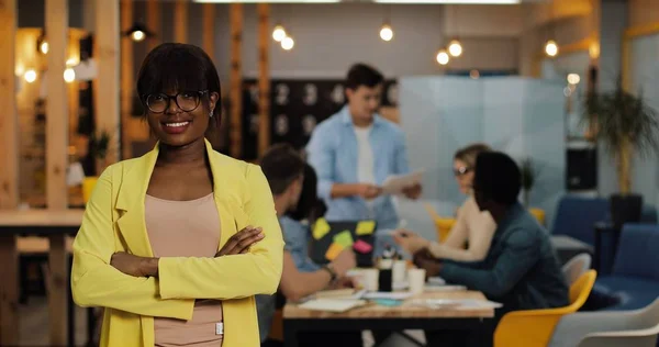 Afro-Amerikalı iş kadını modern ofiste ayakta, gülümseyerek ve kameraya bakıyor. Bulanık arka plan üzerinde çalışan insanlar.