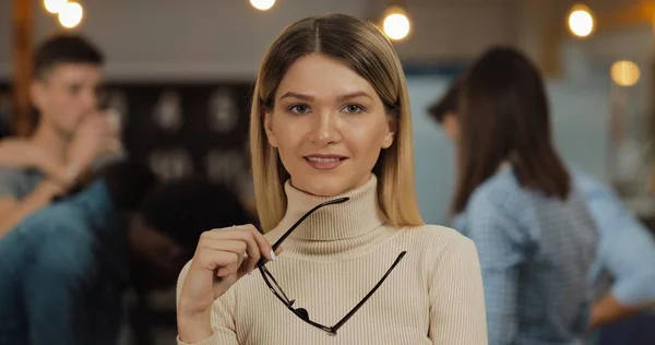 Ofis çalışma alanında duran kameraya bakan şık gözlüklü genç başarılı iş kadınının portresi. Bulanık arka plan üzerinde çalışan insanlar.