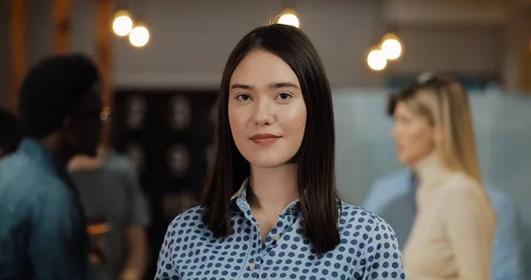 Ofis çalışma alanında duran kameraya bakan genç başarılı iş kadını portresi. Bulanık arka plan üzerinde çalışan insanlar.