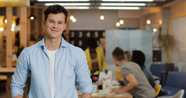 Modern ofiste duran genç gülümseyen başarılı işadamıportresi. Bulanık arka plan üzerinde çalışan insanlar.