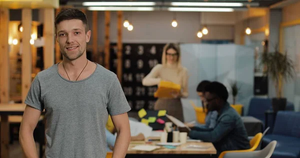 Modern ofiste duran genç gülümseyen başarılı işadamıportresi. Bulanık arka plan üzerinde çalışan insanlar.