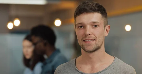 Modern ofiste duran genç gülümseyen başarılı işadamıportresi. Bulanık arka plan üzerinde çalışan insanlar.