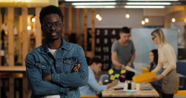 Portrait of young black smiling successful businessman wearing stylish glasses standing in modern office. Working people on the blurred background.