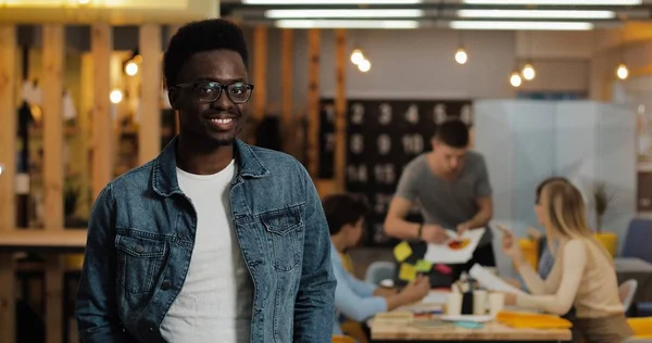 Modern ofiste duran şık gözlük takan genç siyah gülümseyen başarılı işadamı portresi. Bulanık arka plan üzerinde çalışan insanlar.