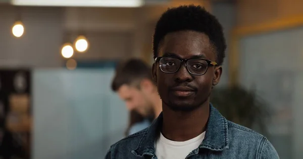 Modern ofiste duran şık gözlük takan genç siyah gülümseyen başarılı işadamı portresi. Bulanık arka plan üzerinde çalışan insanlar.