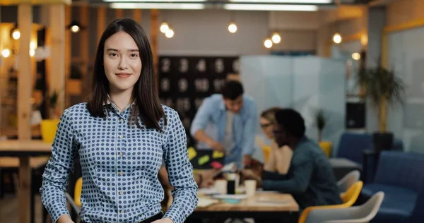 Ofis çalışma alanında duran kameraya bakan genç başarılı iş kadını portresi. Bulanık arka plan üzerinde çalışan insanlar.