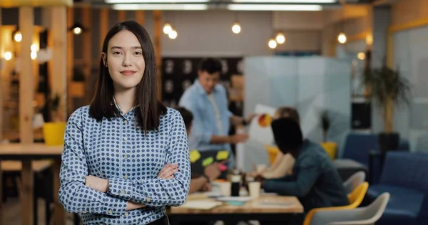 Ofis çalışma alanında duran kameraya bakan genç başarılı iş kadını portresi. Bulanık arka plan üzerinde çalışan insanlar.